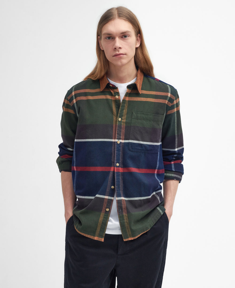 A person with long hair stands against a plain background wearing a Barbour M Bushlane Oversized LS Tartan Shirt in green, blue, red, and white plaid and black pants, with hands in pockets.