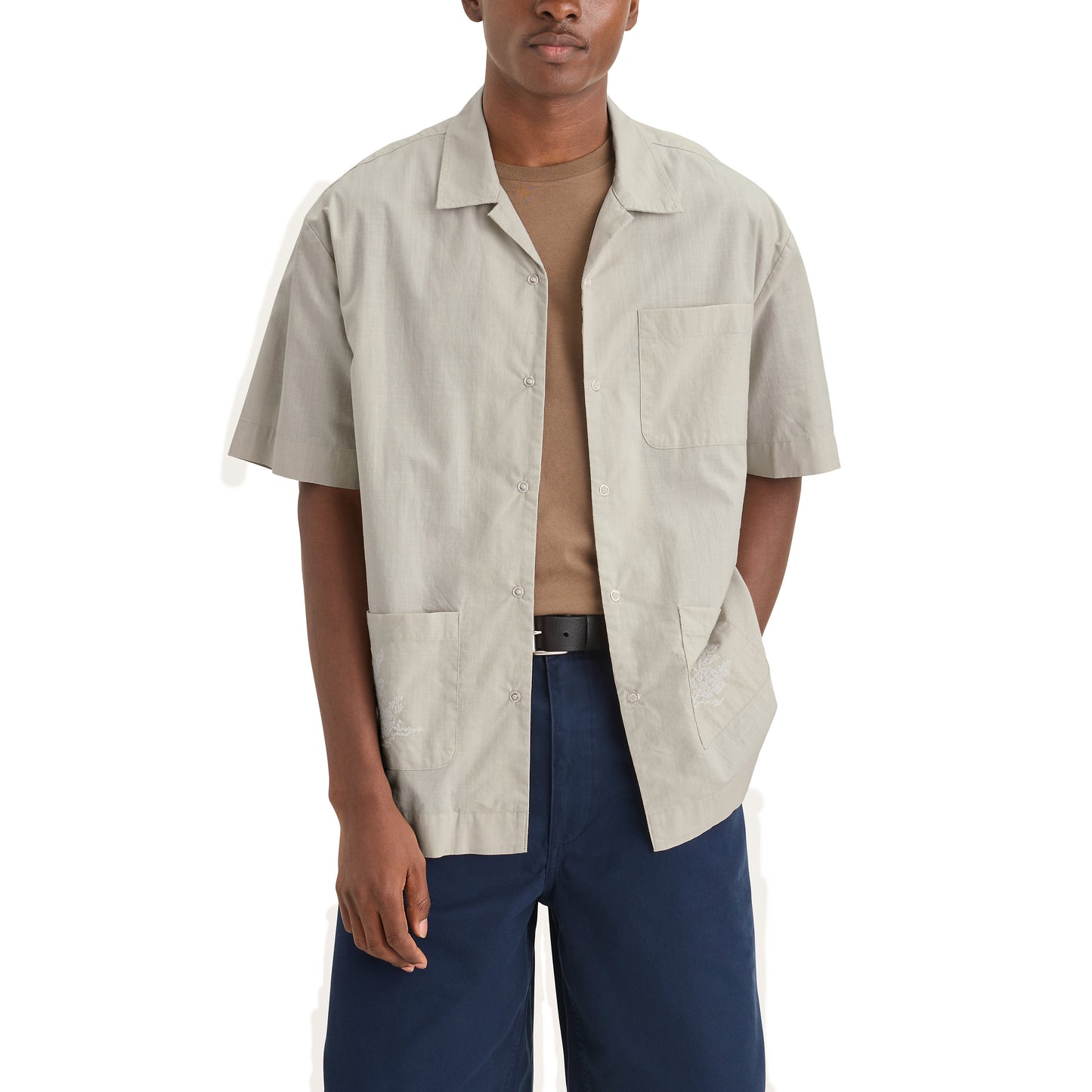 A person stands against a white backdrop, wearing Dockers' Genderless 86 3 Pocket Shirt in light gray over a beige t-shirt with navy pants. The shirt is short-sleeved, it's made of 100% cotton, and it features a camp collar.