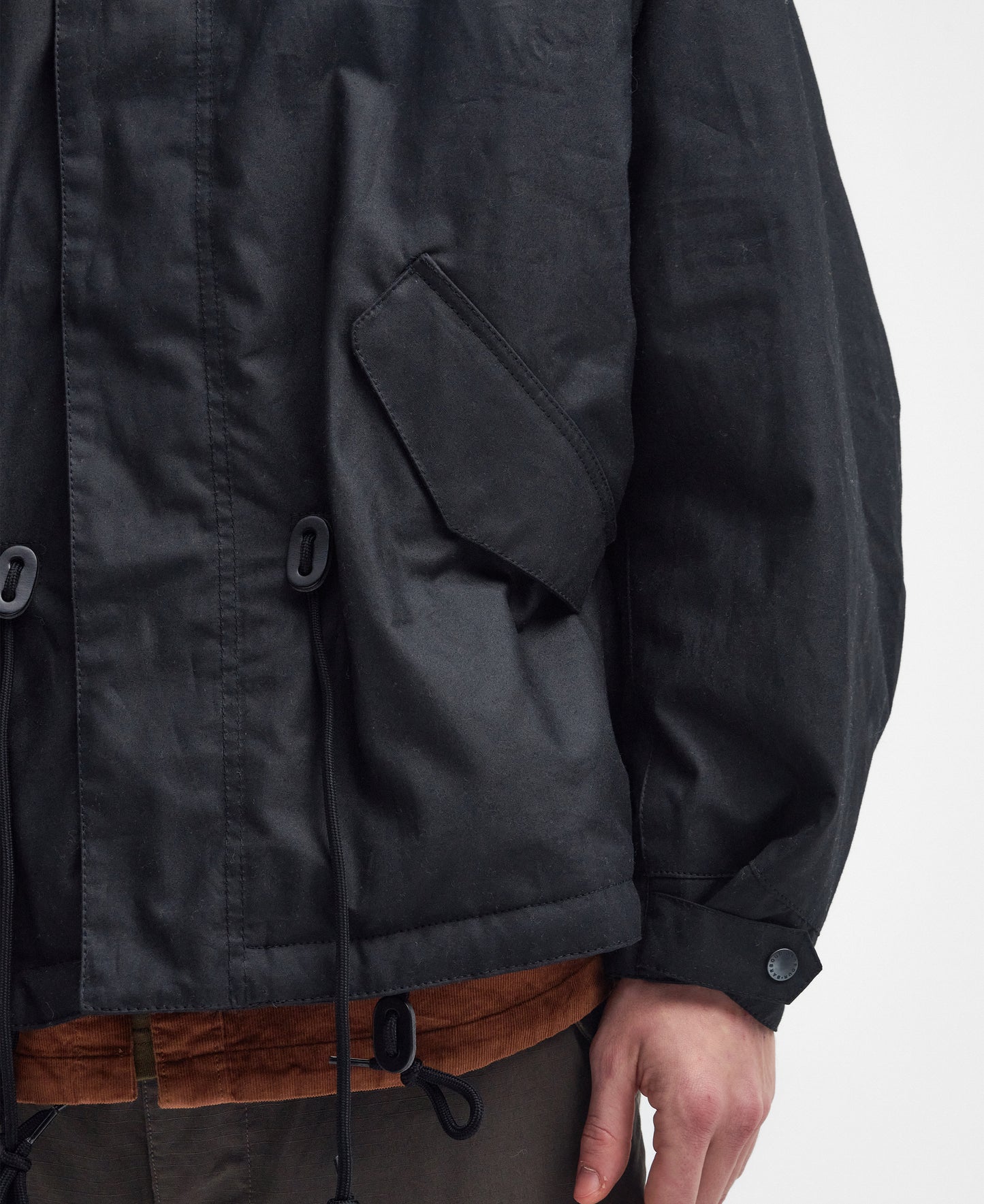 Close-up of a person wearing a dark Barbour Short Wind Waxed Parka featuring drawstrings and a visible pocket flap. Crafted from 6oz waxed cotton, it’s perfect for outdoor adventures. The person is also wearing contrasting shirt and pants, completing the rugged, stylish look.