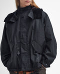 A person is wearing the Barbour Short Wind Waxed Parka in black, crafted from 6oz waxed cotton. The parka features large front pockets and a high collar, as they stand against a plain background. It's perfect for outdoor adventures.