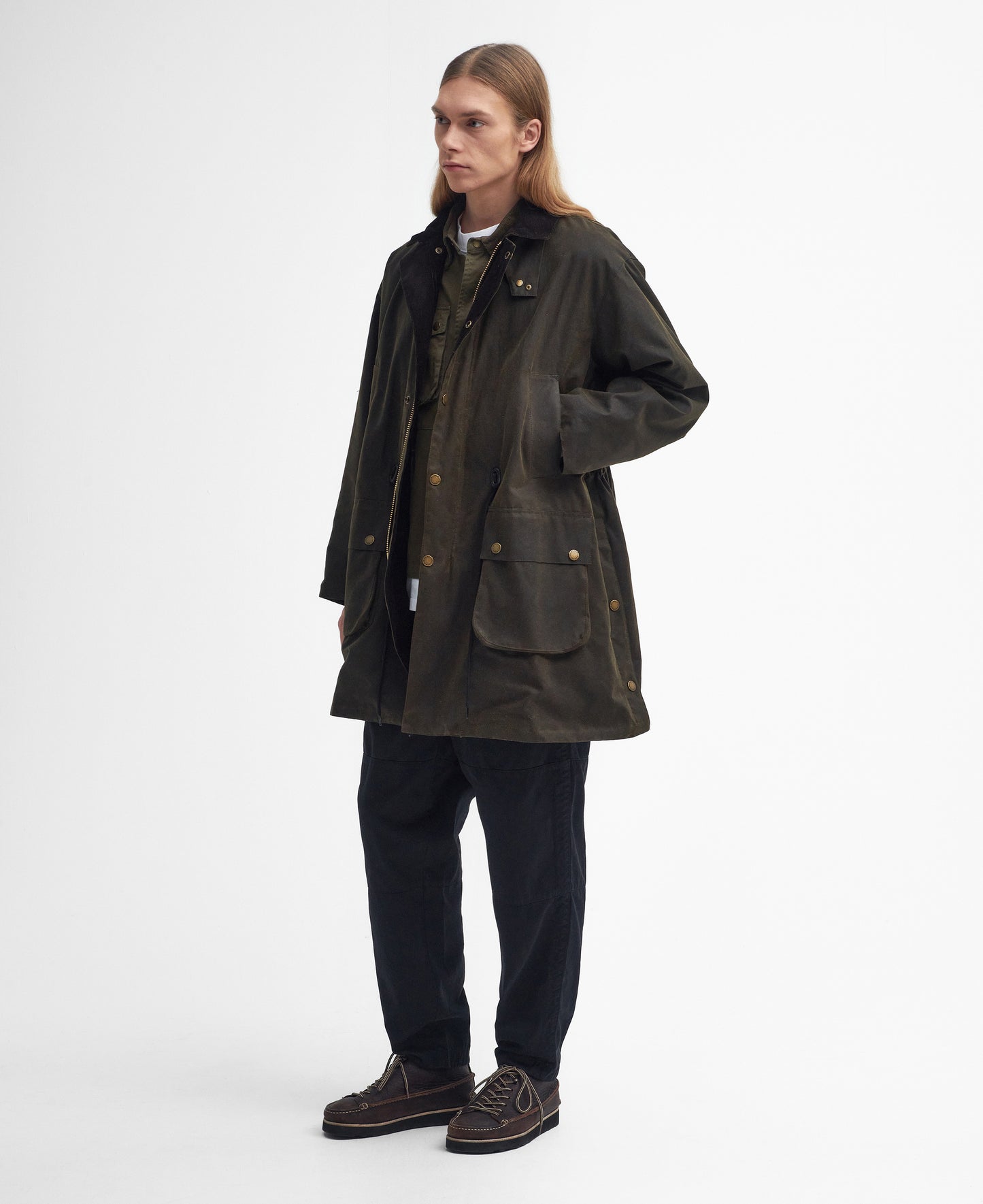 A person with long hair stands modeling a dark green, knee-length M Bedale Wax Trench Coat by Barbour over a green top and black pants. They are wearing dark shoes and looking to the right, exuding a classic look.