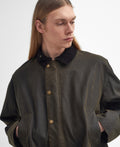 A person with long hair wearing a dark Barbour M Bedale Wax Trench Coat with tartan lining looks to their right with hands in the pockets, exuding a classic look.