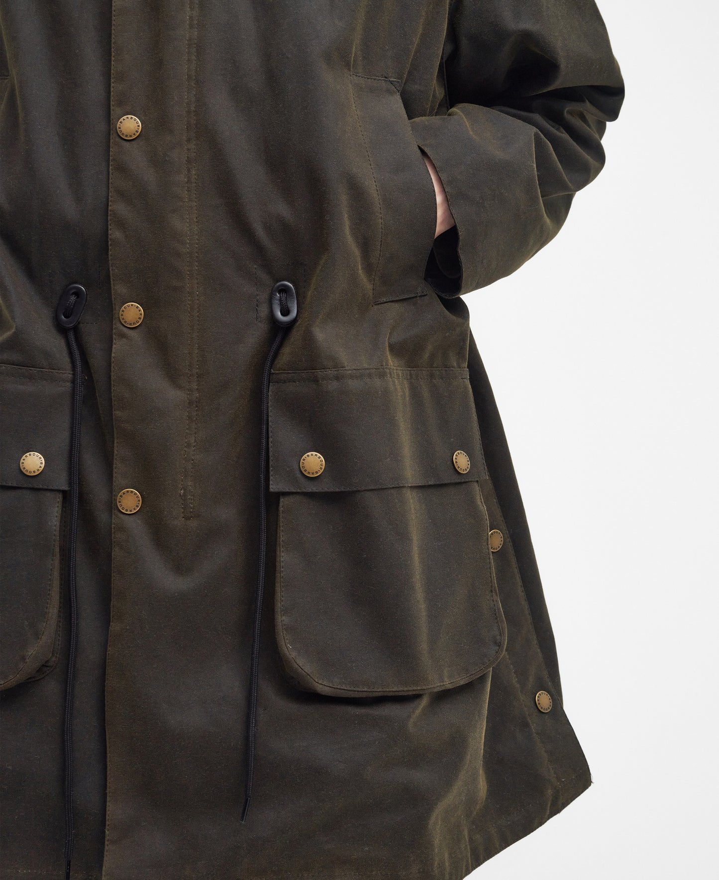 Close-up view of a person wearing a dark green M Bedale Wax Trench Coat by Barbour, featuring large pockets, snap buttons, and black drawstrings, with hands in pockets.