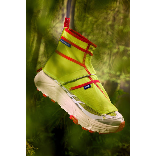 A single Mafate THREE2 Nicole McLaughlin, White/Neon Yellow athletic shoe by Hoka with red accents, branded tags, and a gaiter system, seemingly suspended in mid-air, surrounded by green foliage.