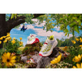 A pair of white and neon yellow Hoka Mafate THREE2 Nicole McLaughlin running shoes displayed outdoors on a tree stump surrounded by yellow flowers, mushrooms, and greenery with a blue butterfly flying nearby. The shoes feature sustainable designs perfect for eco-conscious runners.