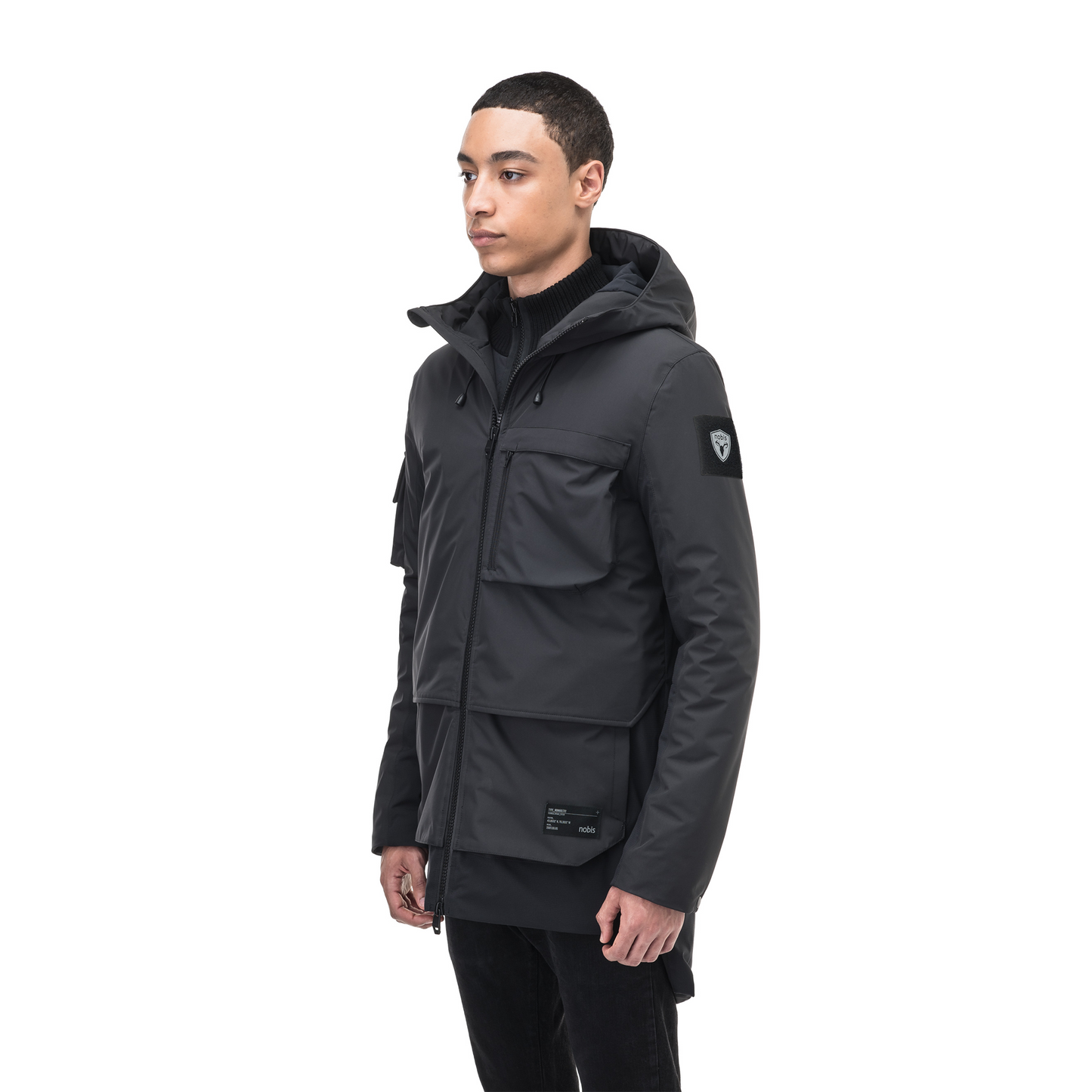 A person stands against a white background wearing the Alta Men's Performance Shell Jacket by Nobis, featuring a dark hood with multiple pockets, Primaloft Gold Insulation, and a Durable Water Repellent finish.