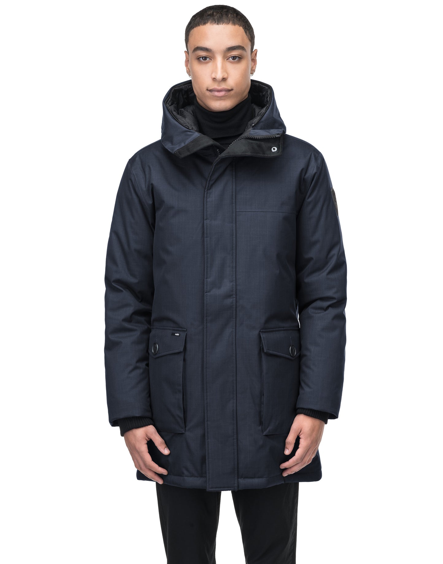 A person wearing the Yves Men's Parka by Nobis—a stylish, dark blue winter coat featuring a hood and front pockets—stands against a plain white background. The slim-fitting jacket is perfect for winter conditions.