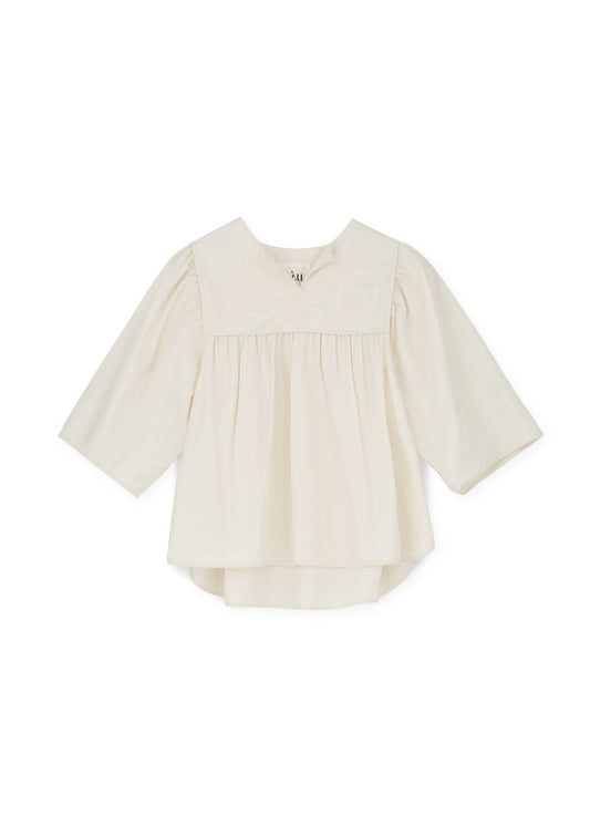 The Nomi Blouse by Aiayu is a cream-colored, long-sleeved blouse with a rounded neckline and loose fit, featuring pleated detailing on the front, made from regenerative cotton.