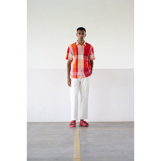 A man stands against a minimalistic background wearing the Lax Short Sleeve Shirt in Red Multi from Original Madras Trading Co., white pants, and red sandals.