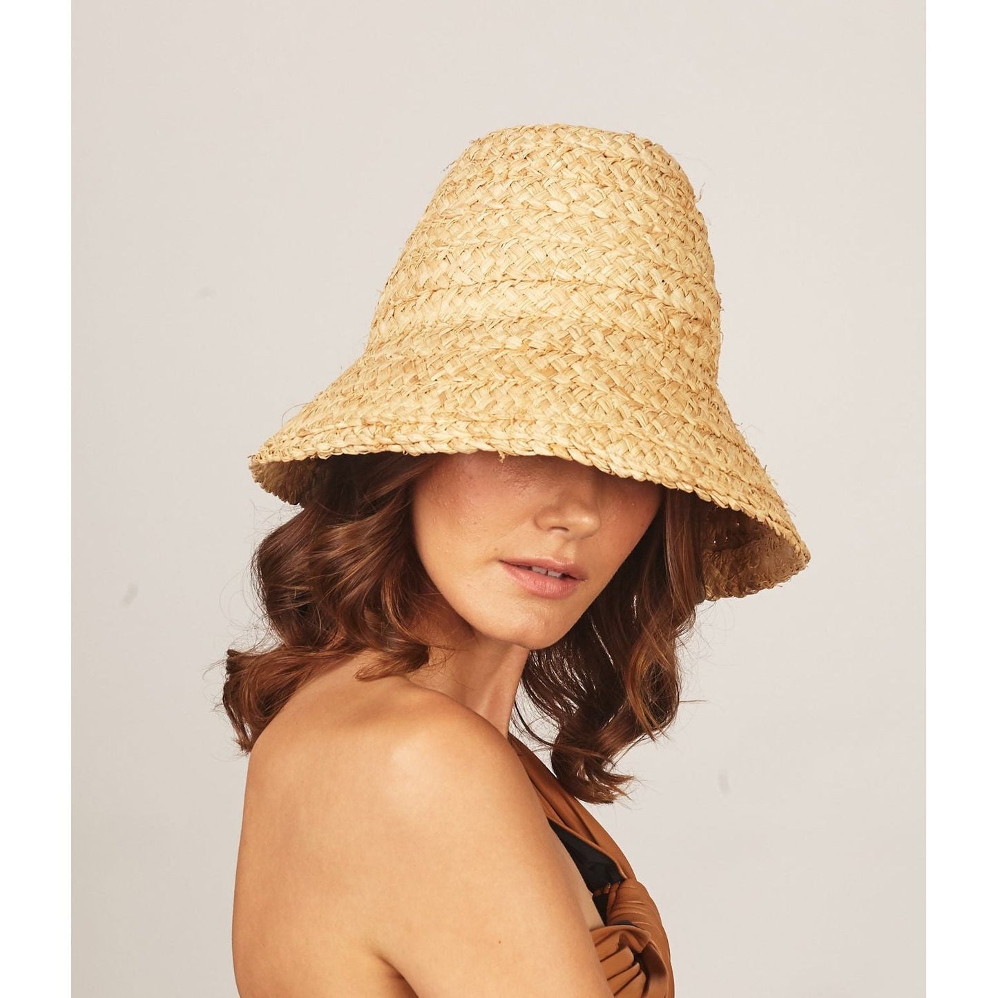 A person wearing a Lola Hats Petit Nap Hat, Natural made from natural raffia braid that partially obscures their face, with long brown hair and a light-colored, strapless top.