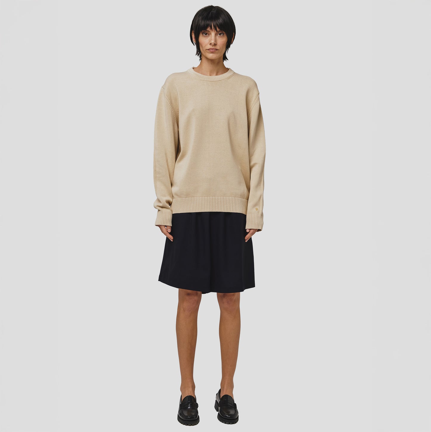 A person models the Seven Gauge Cotton Crewneck Sweater in beige, paired with a black skirt and shoes, on a plain white background, highlighting the elegance of this knitwear ensemble.