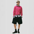 A person wears Seven Gauge's Cotton Crewneck Sweater in pink over a gray organic cotton shirt, paired with black knee-length shorts, socks, and loafers, confidently posing against a plain background.