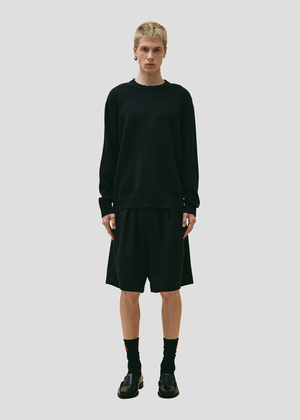 A person wearing a black Cotton Crewneck Sweater from Seven Gauge, along with black shorts, black socks, and black shoes, stands against a plain gray background. The sleek design of the knitwear enhances the minimalist setting with subtle sophistication.