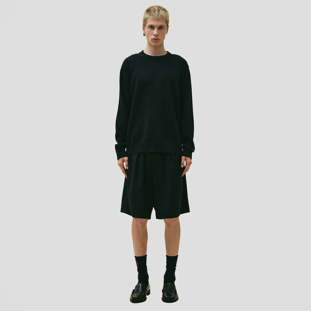 A person stands against a plain background, wearing Seven Gauge's black Cotton Crewneck Sweater made from organic cotton, paired with knee-length black shorts. The look is completed with black socks and shoes, embodying the refined quality of 7-gauge knitwear.