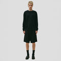 A person stands against a plain background, wearing Seven Gauge's black Cotton Crewneck Sweater made from organic cotton, paired with knee-length black shorts. The look is completed with black socks and shoes, embodying the refined quality of 7-gauge knitwear.