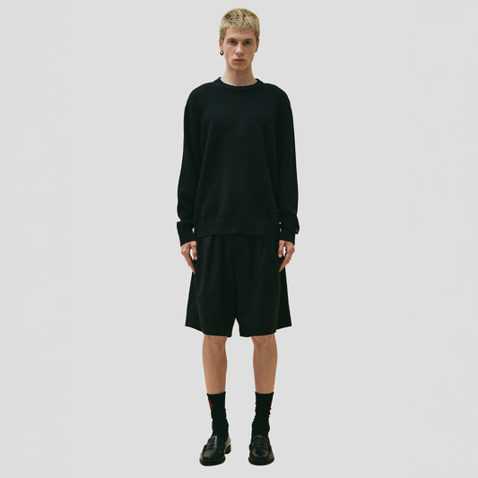 A person stands against a plain background, wearing Seven Gauge's black Cotton Crewneck Sweater made from organic cotton, paired with knee-length black shorts. The look is completed with black socks and shoes, embodying the refined quality of 7-gauge knitwear.