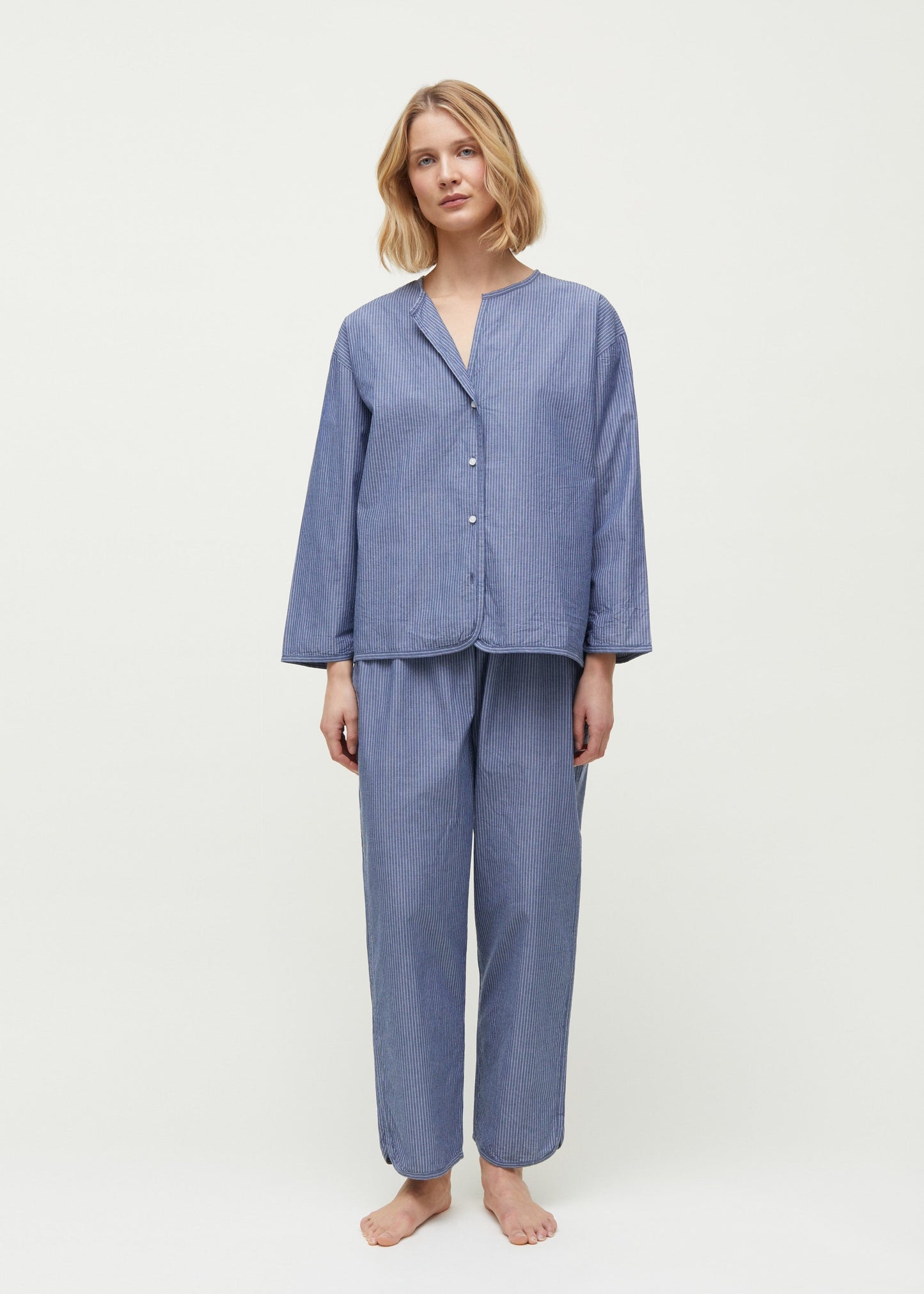 A woman stands barefoot, wearing a loose-fitting, long-sleeve pajama set made from Chetna Organic Cotton by Aiayu, featuring the Letto Casual Pant and a button-up top. She has shoulder-length blonde hair and a neutral expression. The background is plain white.