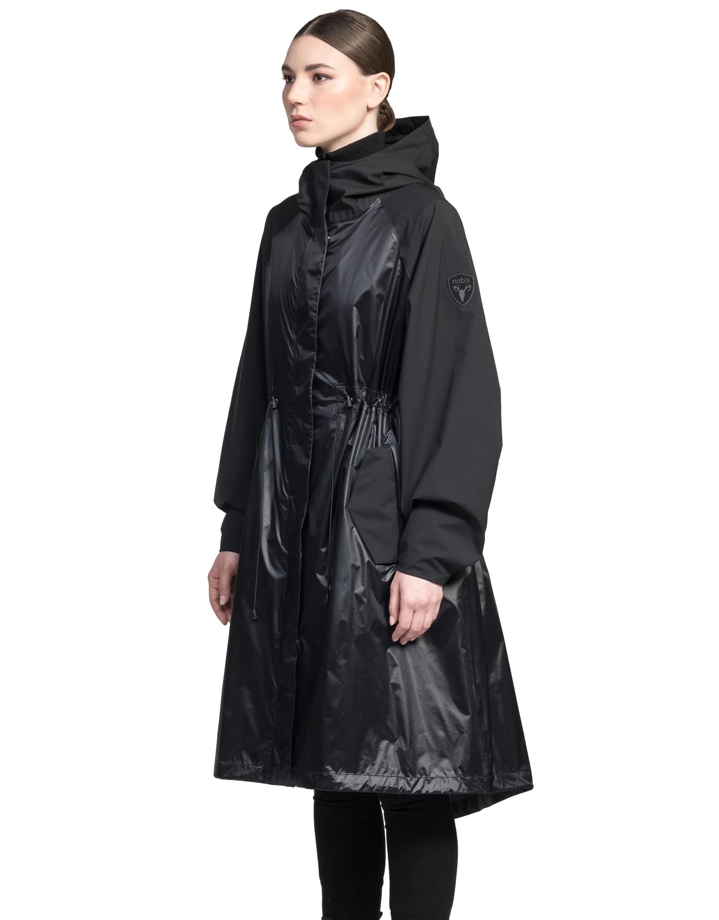 A person stands against a plain white background, wearing the Reyna Women's Packable Long Shell Jacket by Nobis. This black hooded coat has a shiny DWR coating and features adjustable drawcords for a customizable fit.