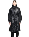 A person is standing against a plain background wearing the Reyna Women's Packable Long Shell Jacket by Nobis, featuring a black hooded design with adjustable drawcords at the cinched waist.