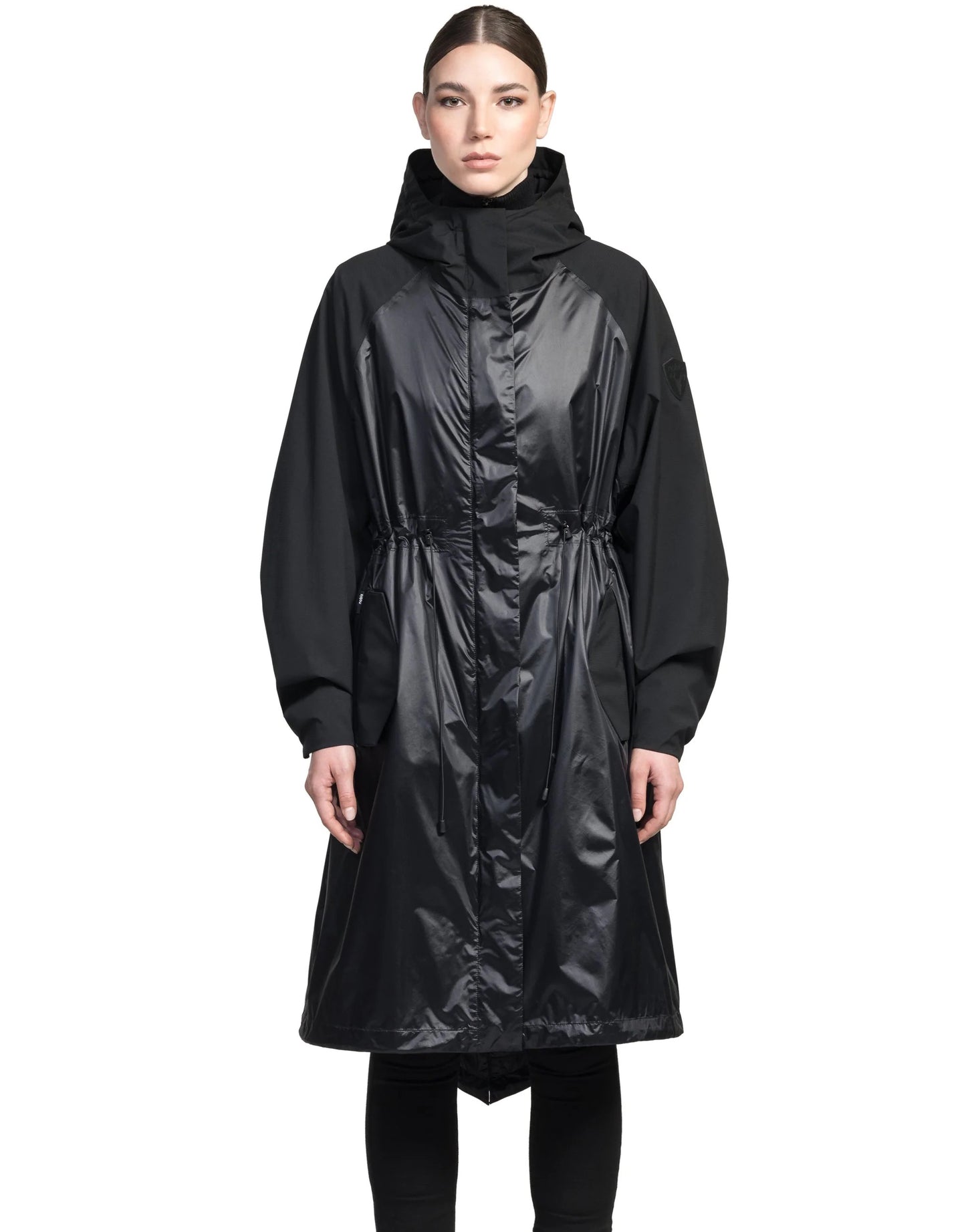 A person is standing against a plain background wearing the Reyna Women's Packable Long Shell Jacket by Nobis, featuring a black hooded design with adjustable drawcords at the cinched waist.