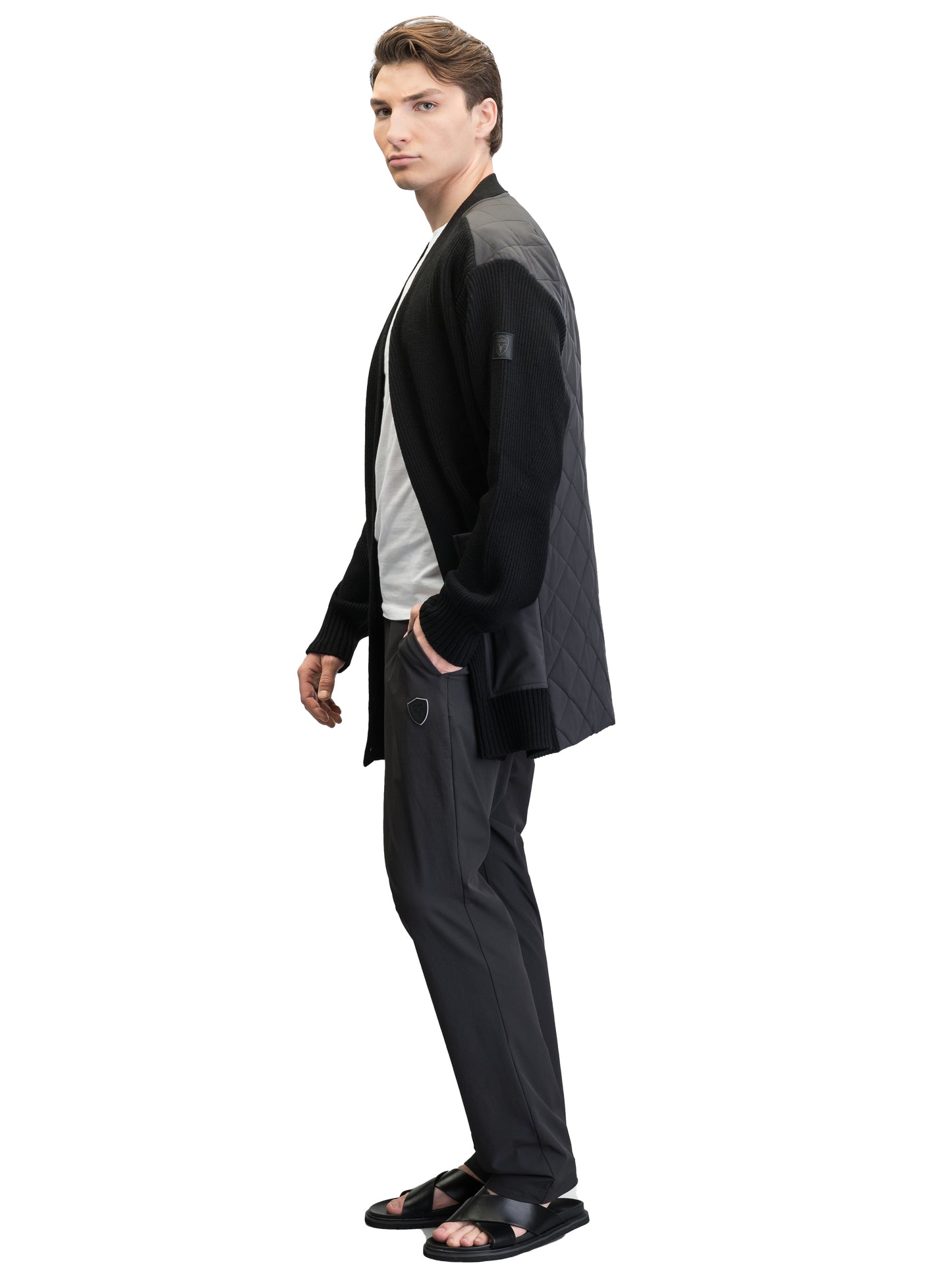 A person stands wearing a black Riga Tailored Button Front Cardigan by Nobis, known for its durability, paired with a white shirt, dark pants, and black sandals against a plain background.