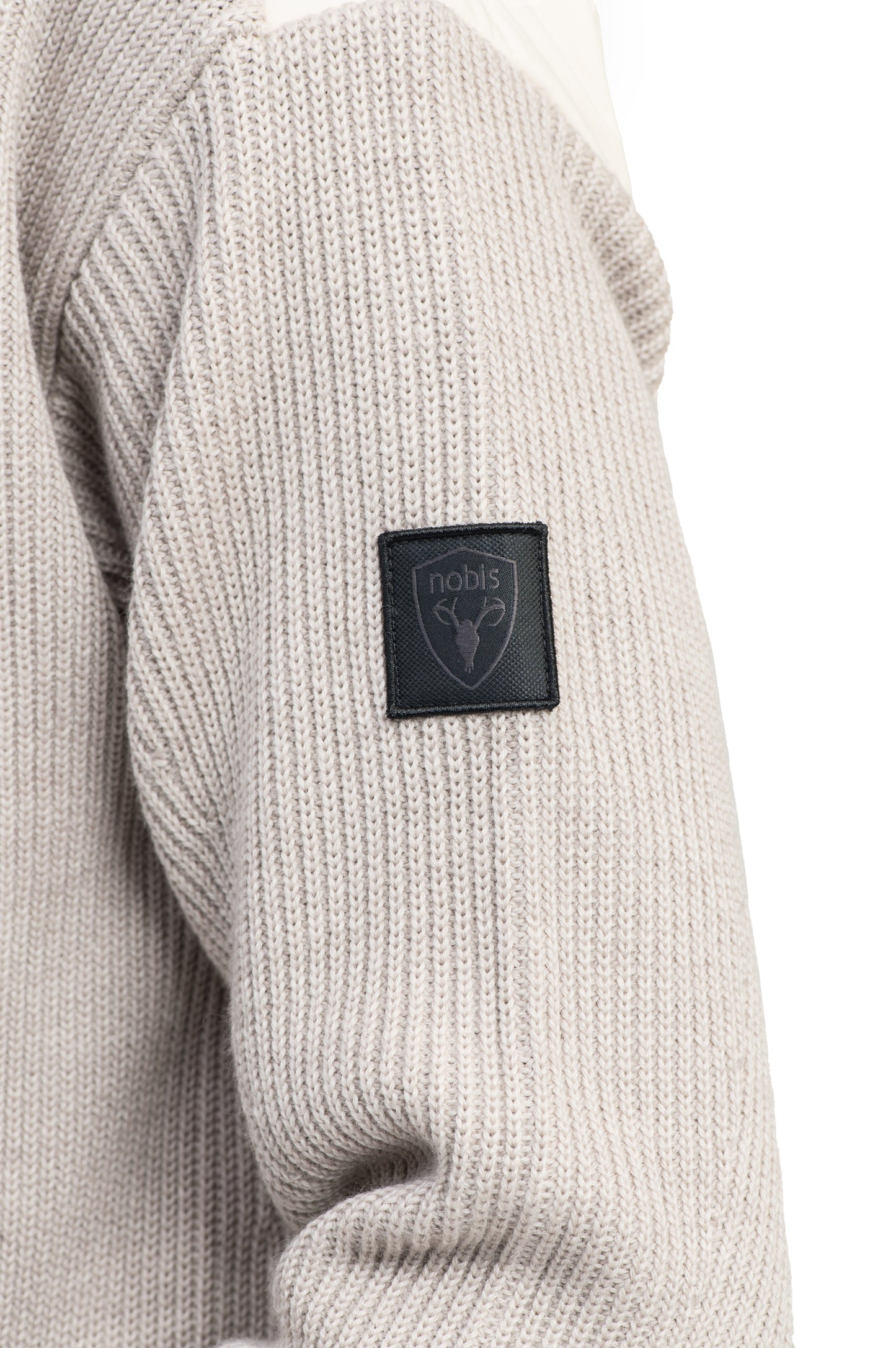 Close-up of a beige knitted sleeve from the Riga Tailored Button Front Cardigan by Nobis, featuring a black patch with the "nobis" branding and an antler logo, showcasing its 100% virgin extra fine merino wool for exceptional durability.