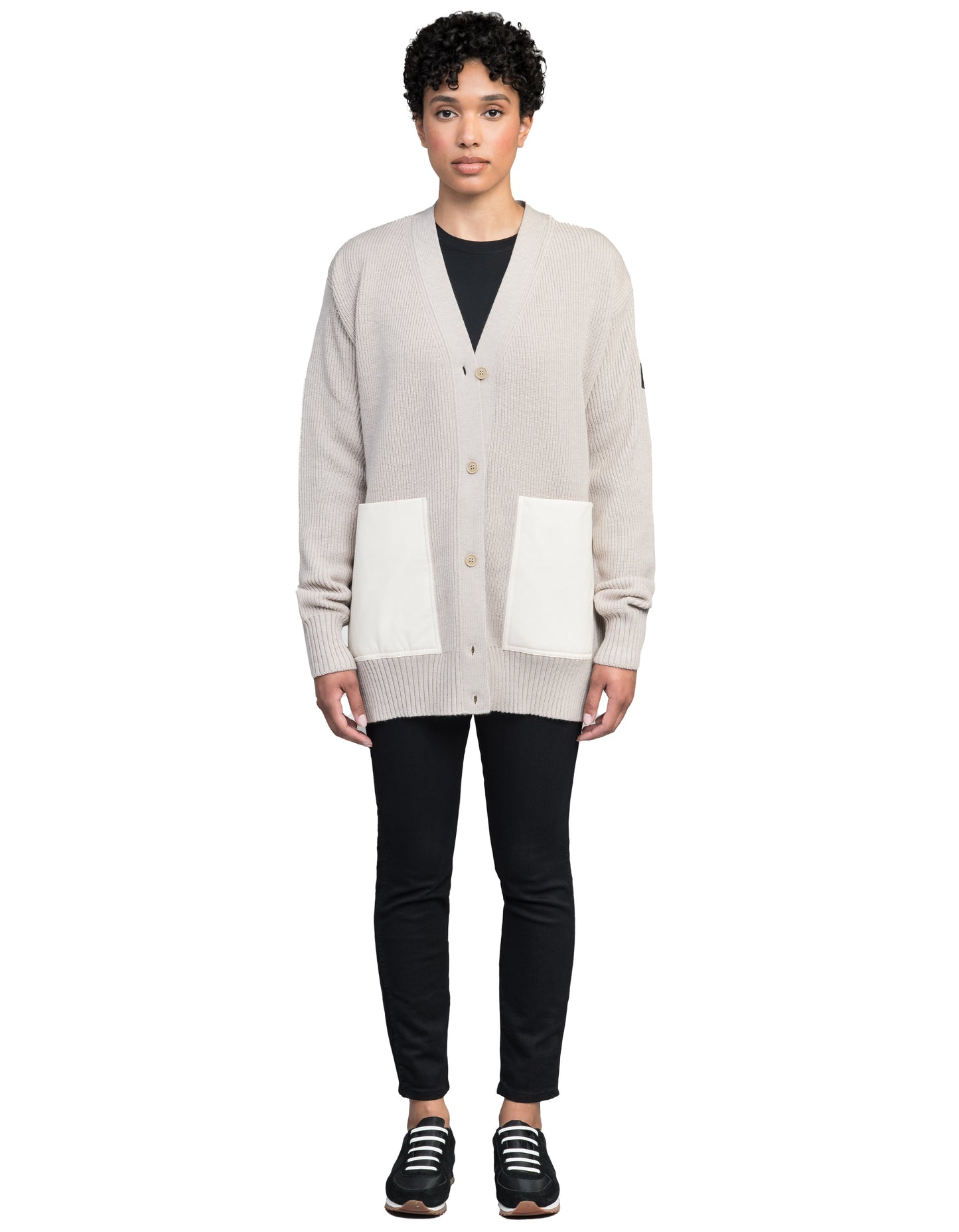 A person dressed in the Riga Tailored Button Front Cardigan by Nobis, crafted from 100% virgin extra fine merino wool and known for its durability, paired with black pants and sneakers, stands against a plain white background.