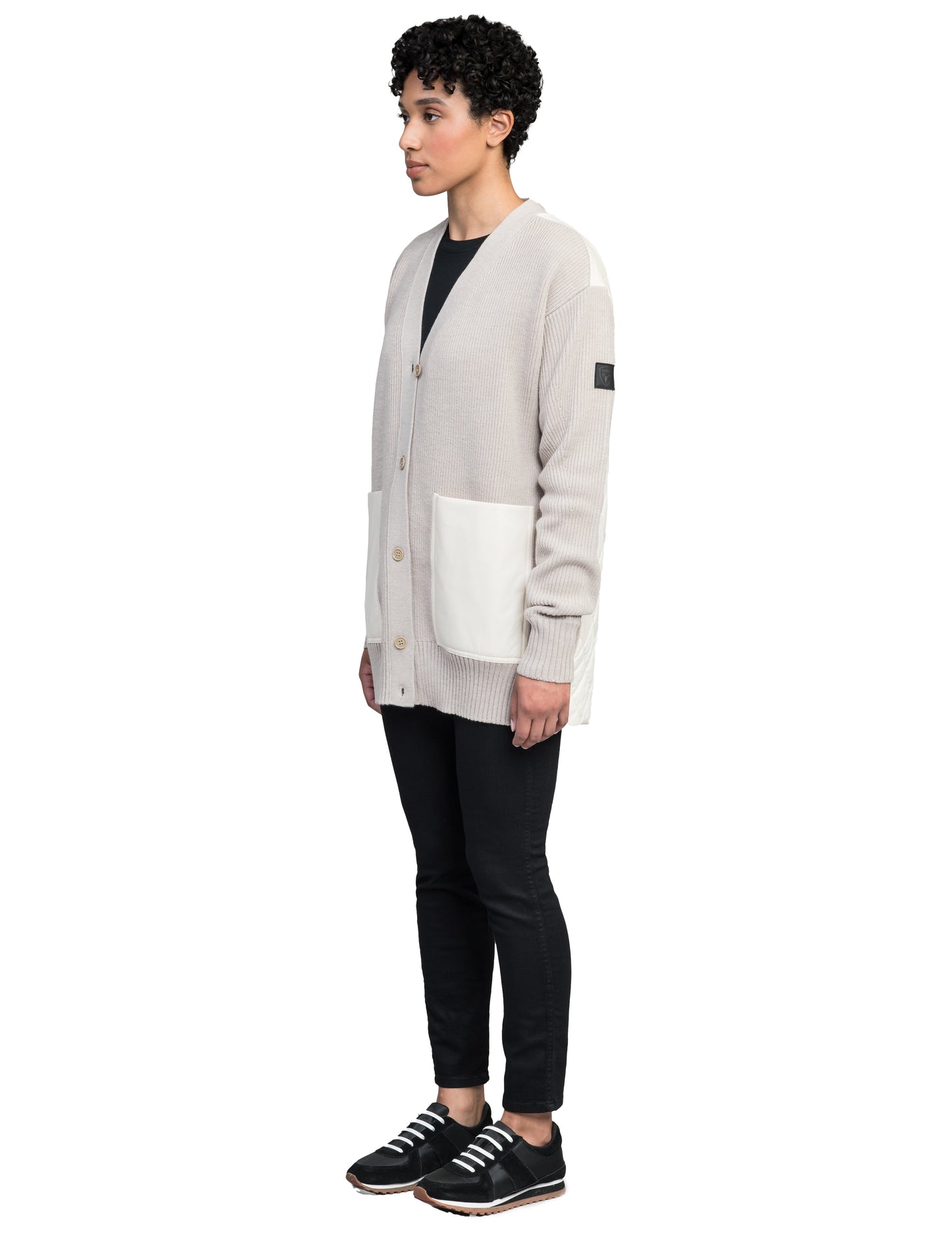 A person stands wearing the Riga Tailored Button Front Cardigan by Nobis, in beige, crafted from 100% virgin extra fine merino wool and featuring white pockets. It's paired with a black shirt, black pants, and black sneakers against a white background.