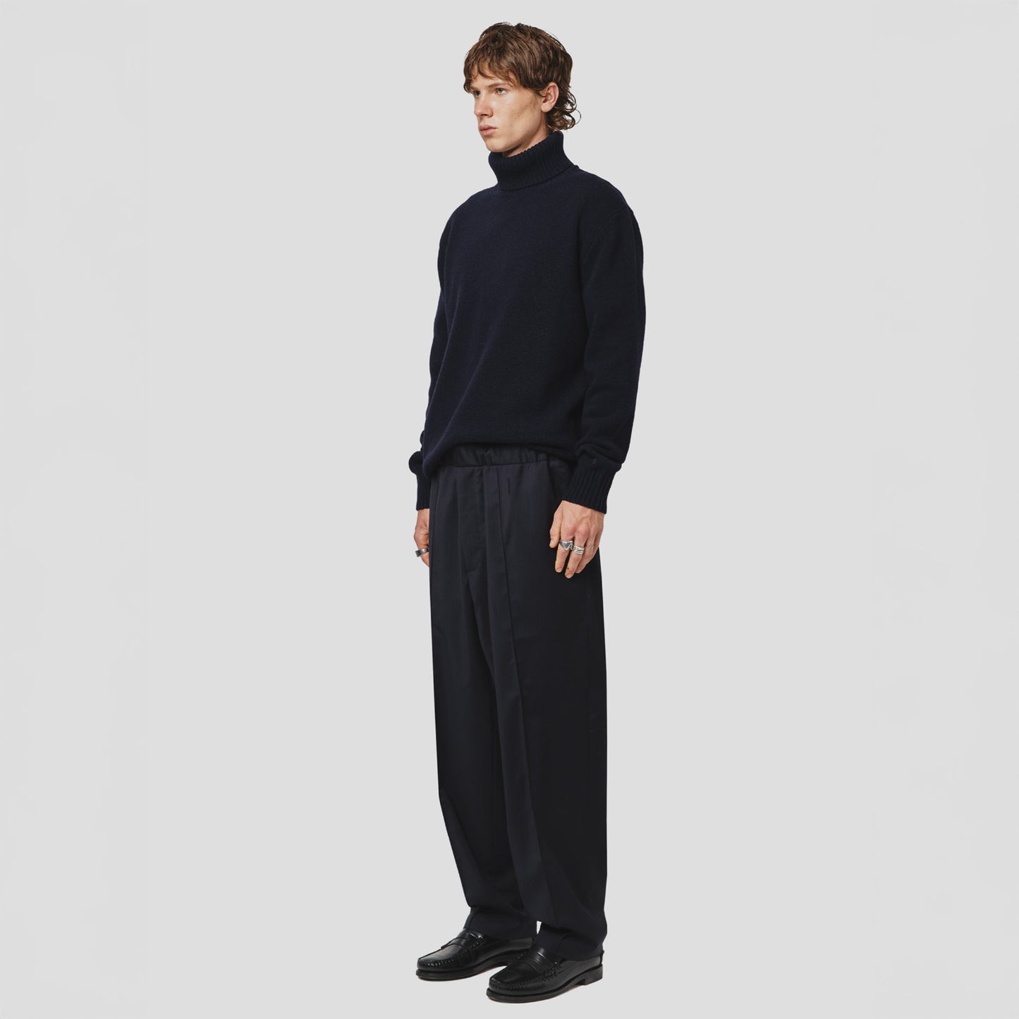 A person stands against a plain background, wearing Seven Gauge's Lambswool High Neck Sweater made from RWS-certified Australian wool, loose-fitting black pants, and black shoes, looking to the side.