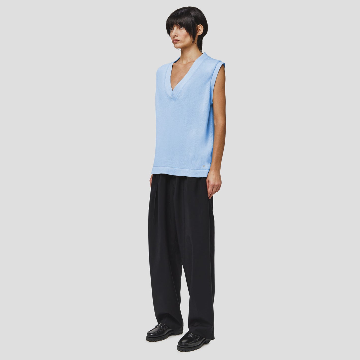 A person stands against a white background, wearing the Seven Gauge Cotton Vest Sweater in light blue with organic cotton, paired with loose black pants and black shoes.