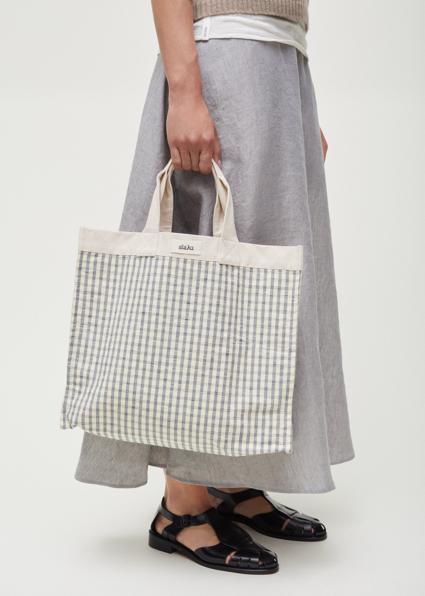 A person holding the Aiayu Shopper Check made of 100% organic Chetna cotton in their left hand, wearing a beige top, grey maxi skirt, and black strappy shoes.