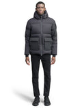 A man dressed in the Supra Men's Performance Puffer by Nobis, featuring a weather-resistant shell and heat-sealing ribbed cuffs, paired with black pants and sneakers, is standing against a white background.