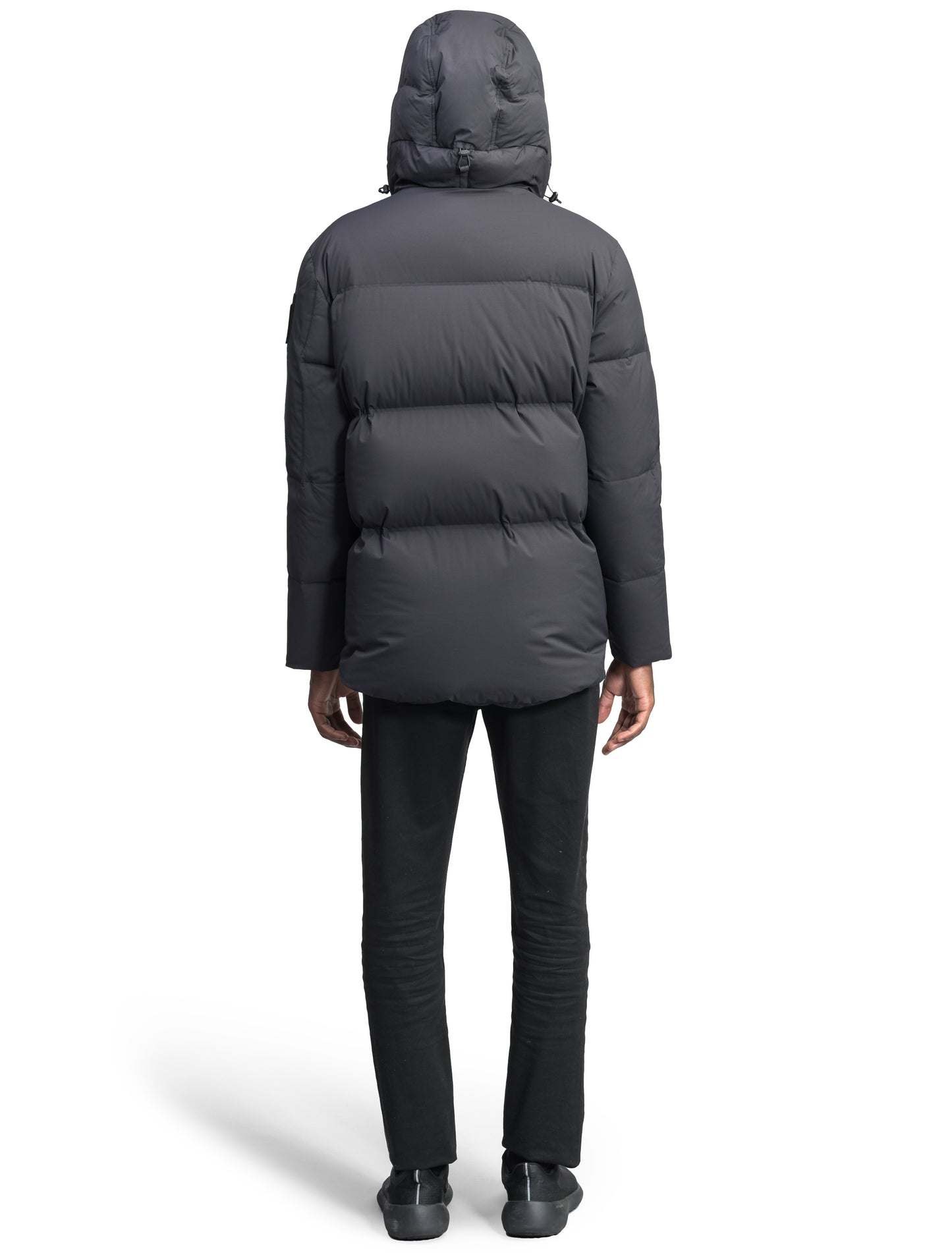 A person wearing the Supra Men's Performance Puffer by Nobis, a dark hooded jacket filled with Premium Canadian White Duck Down and featuring heat-sealing ribbed cuffs, is paired with dark pants and shoes while standing with their back to the camera.