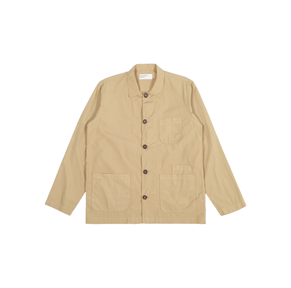 The Universal Works Bakers Overshirt in sand is a beige, breathable cotton jacket with a collar and long sleeves. It has three front pockets, including one on the chest, and features durable dark buttons.