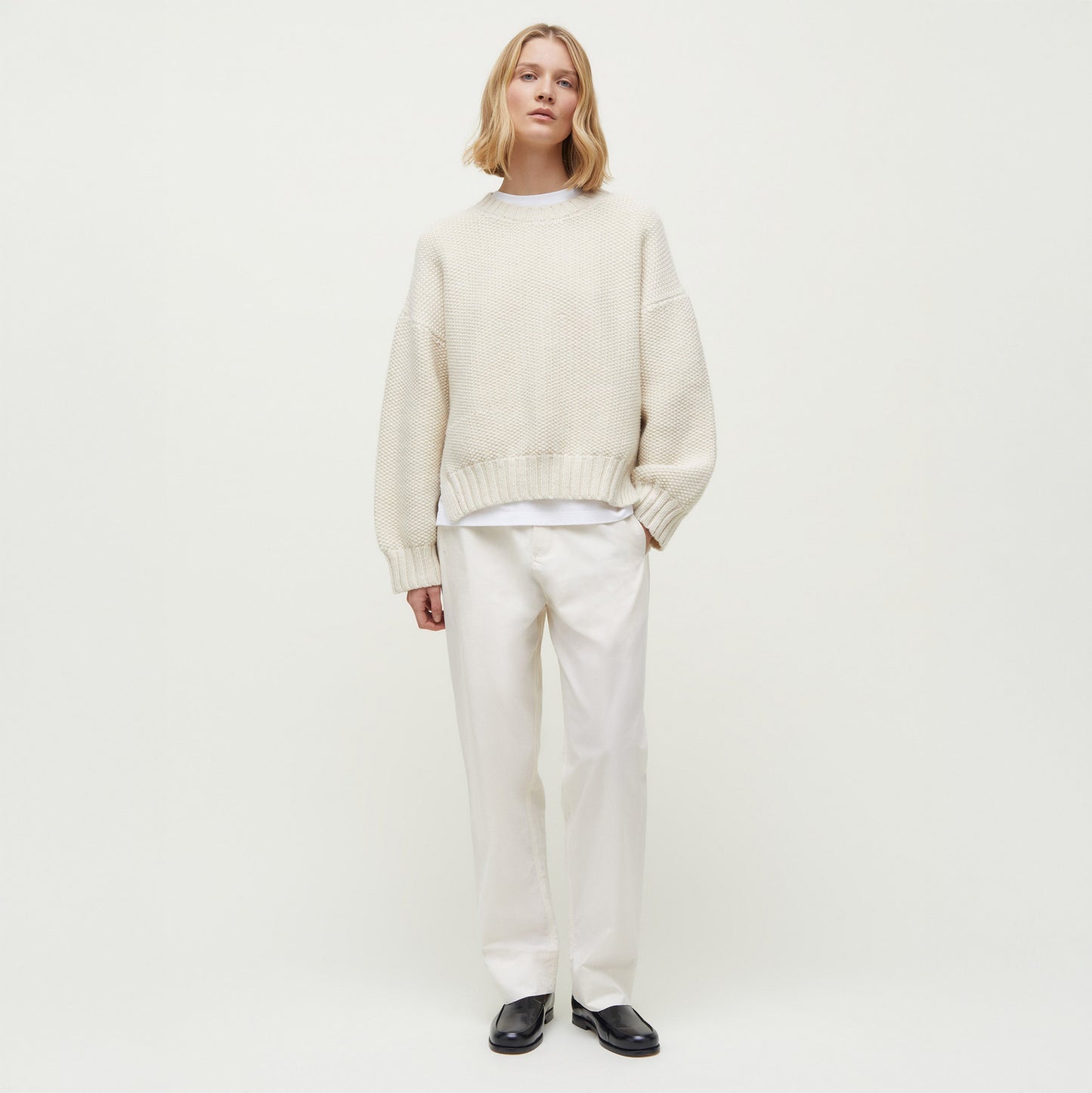 A person wearing an Aiayu Vetla Sweater in cream, paired with white pants and black shoes, stands against a plain white background.