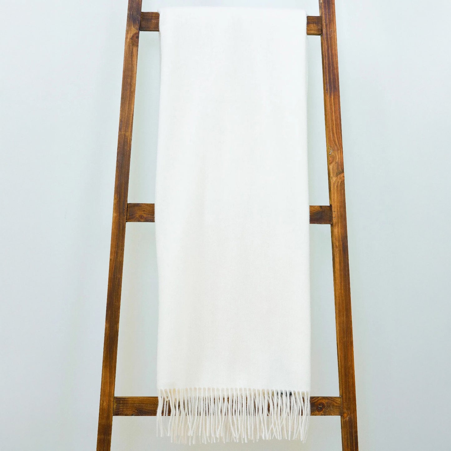 The Classic Throw by Alashan, a white ultrafine merino wool blanket with fringe, is draped over a wooden ladder against a light-colored wall.