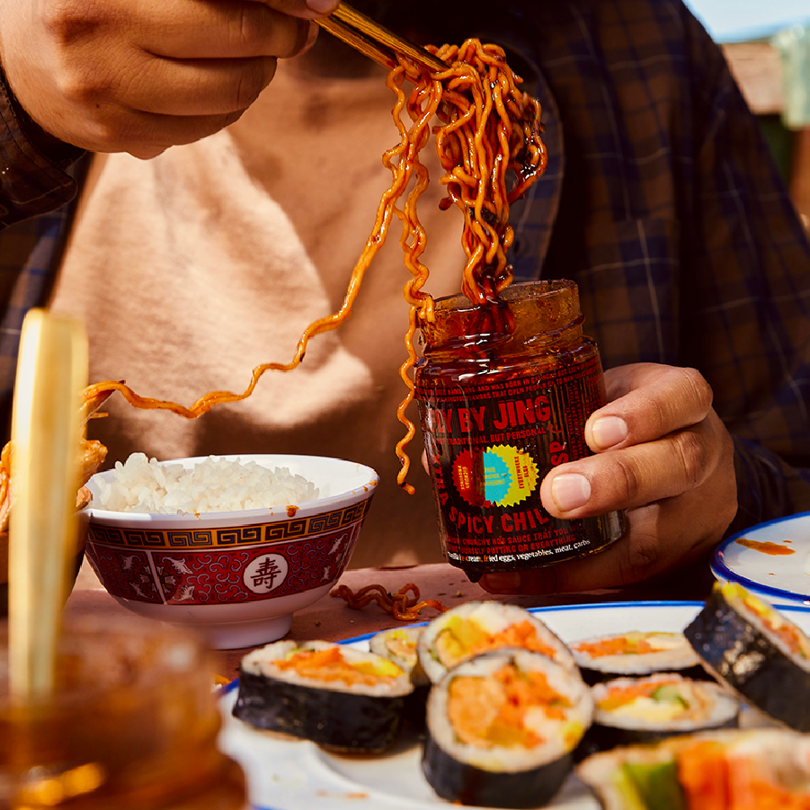 A person uses chopsticks to pick up noodles from a jar of Fly By Jing Xtra Spicy Chili Crisp, with bowls of rice and assorted sushi on a table.