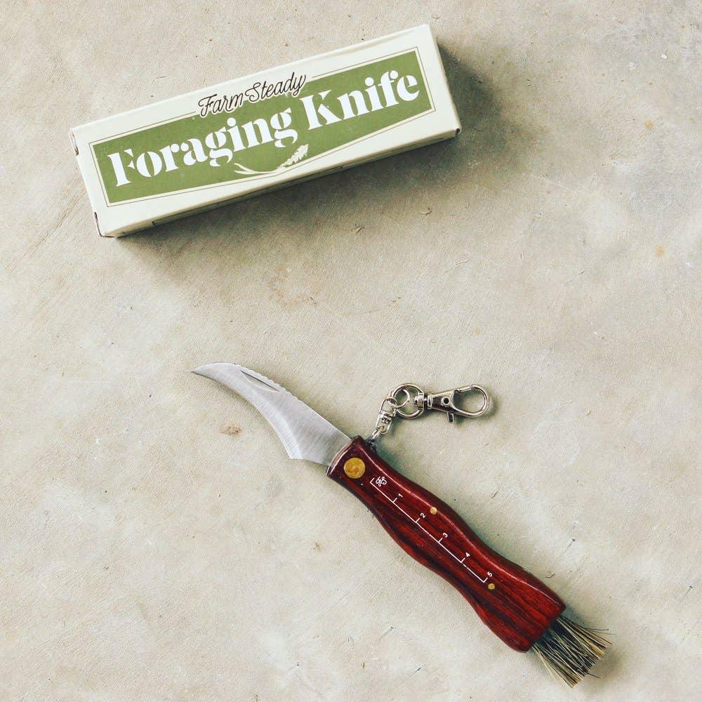 A Foraging Tool Kit with a curved blade and wooden handle, part of a comprehensive foraging kit, lies on a light-colored surface next to a box labeled "FarmSteady Foraging Tool Kit.