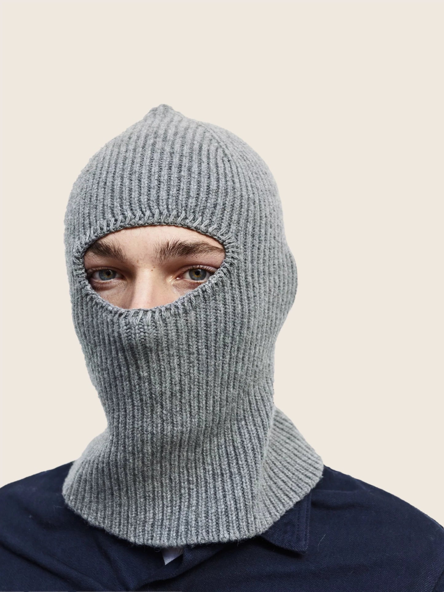 A person wearing a gray knitted balaclava by Le Bonnet, featuring eye and mouth openings and crafted from soft Merino wool, against a plain background.