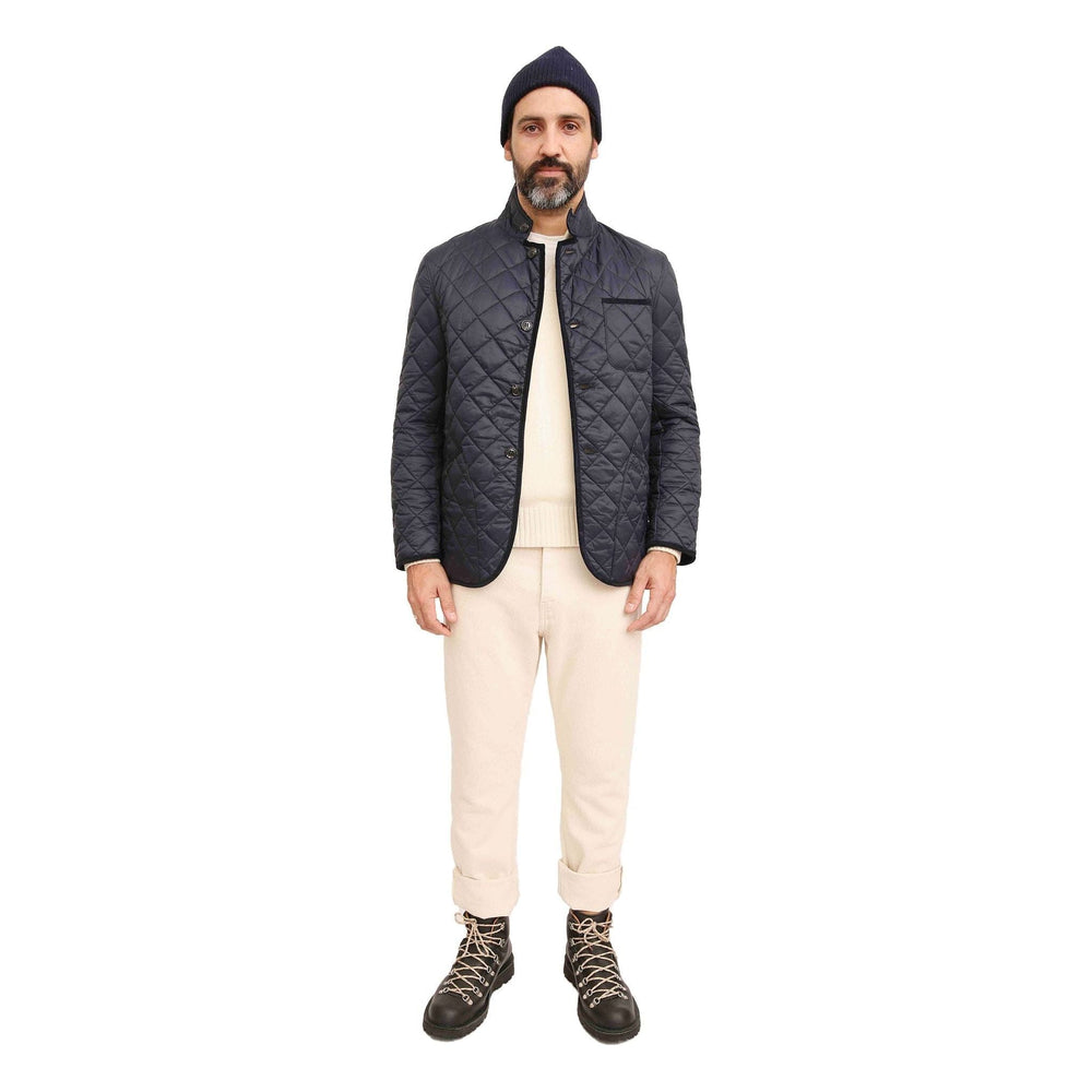 A man stands wearing a navy Men's Modern Liddesdale Quilted Jacket by Barbour featuring a button tab closure and tailored fit, paired with beige pants, a light sweater, a dark beanie, and black boots against a plain white background.