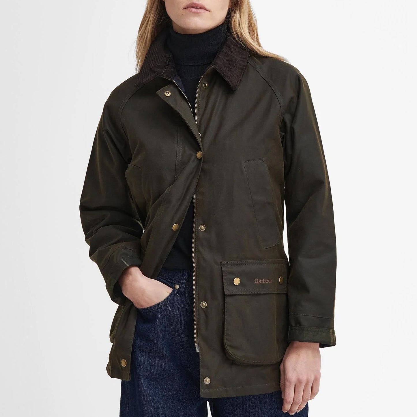 A person stands with hands in pockets, wearing the Barbour Acorn Wax Jacket featuring a corduroy collar and hint of tartan lining, paired with a sleek black turtleneck and classic blue jeans.