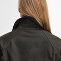 Rear view of a person with long blonde hair wearing the Acorn Wax Jacket by Barbour, showcasing a dark green waxed cotton design with Barbour tartan lining and an upturned collar.