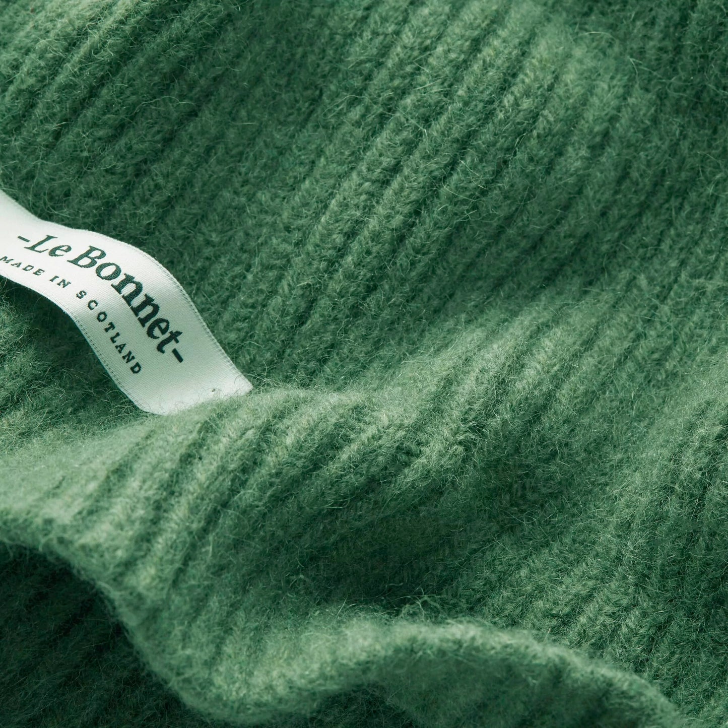 A close-up of a jade ribbed lambswool scarf features a small label reading "Le Bonnet - Made in Scotland." The biodegradable nature of the wool adds an eco-friendly touch to this elegant Le Bonnet piece.