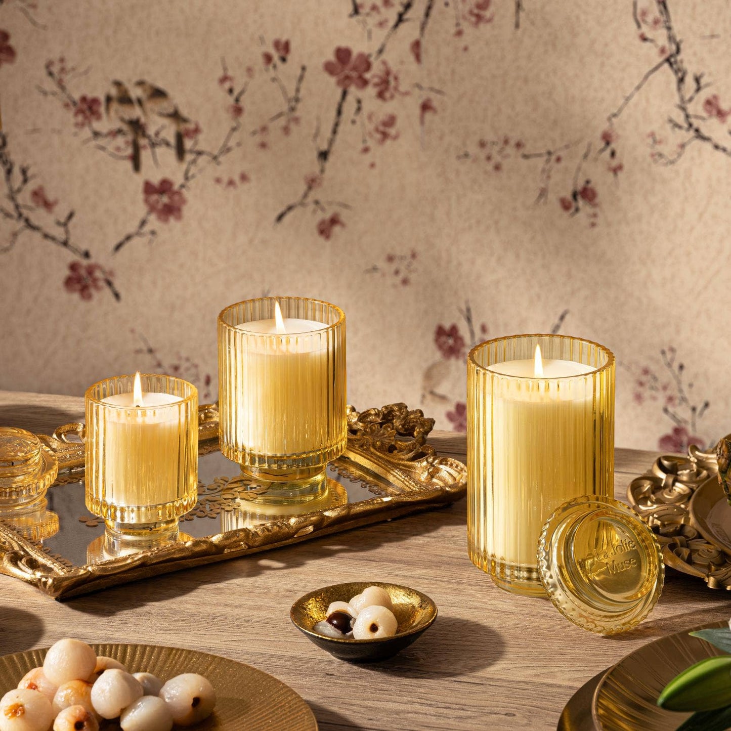 Three Amelie - Tahitian Lychee candles from La Jolie Muse, made with natural soy wax and set on a decorative tray, exude their tropical sweetness amidst plates of lychee and nuts against floral wallpaper.