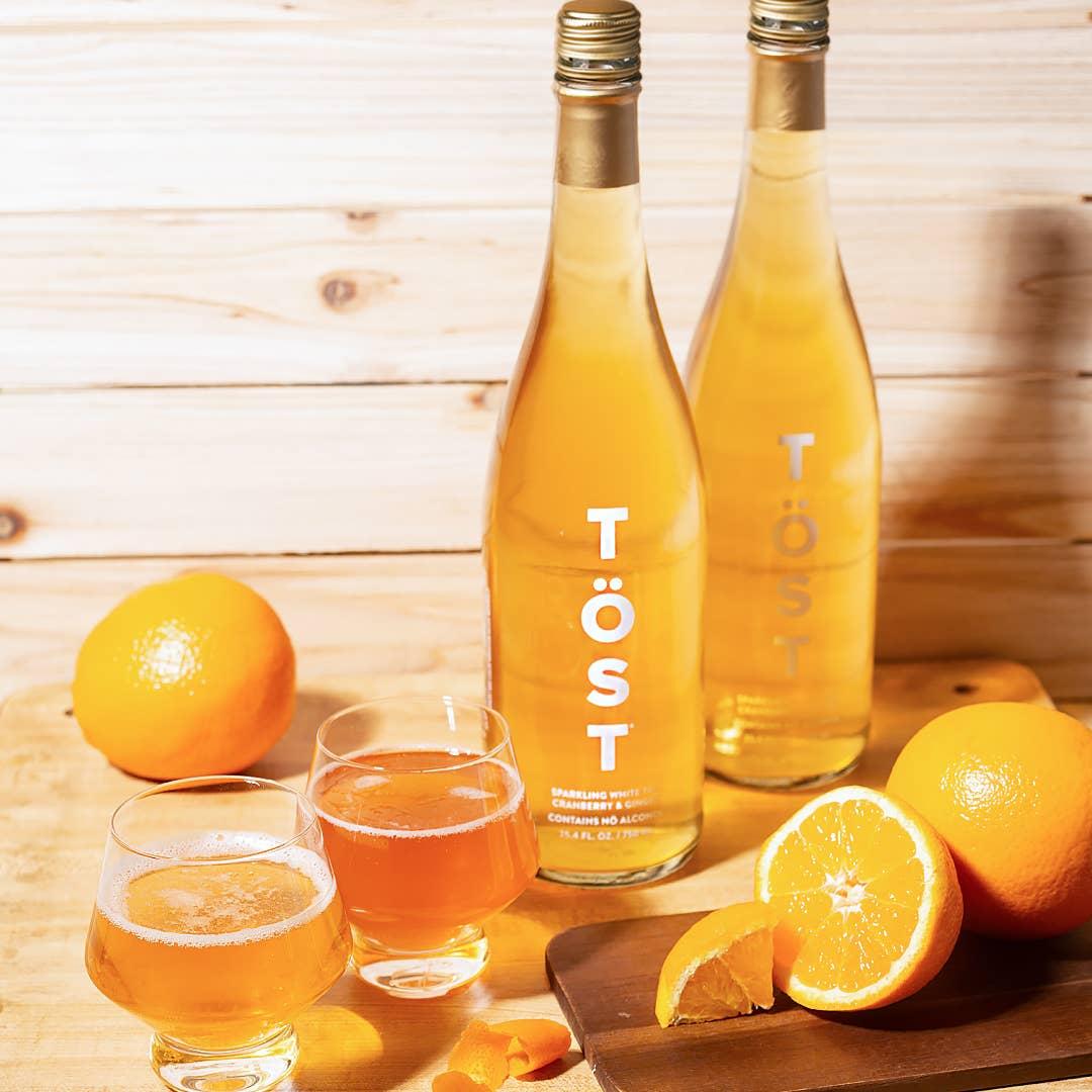 Two bottles of TÖST white tea cranberry ginger non-alcoholic beverage beside glasses half-filled with the drink, and whole and sliced oranges on a wooden surface.