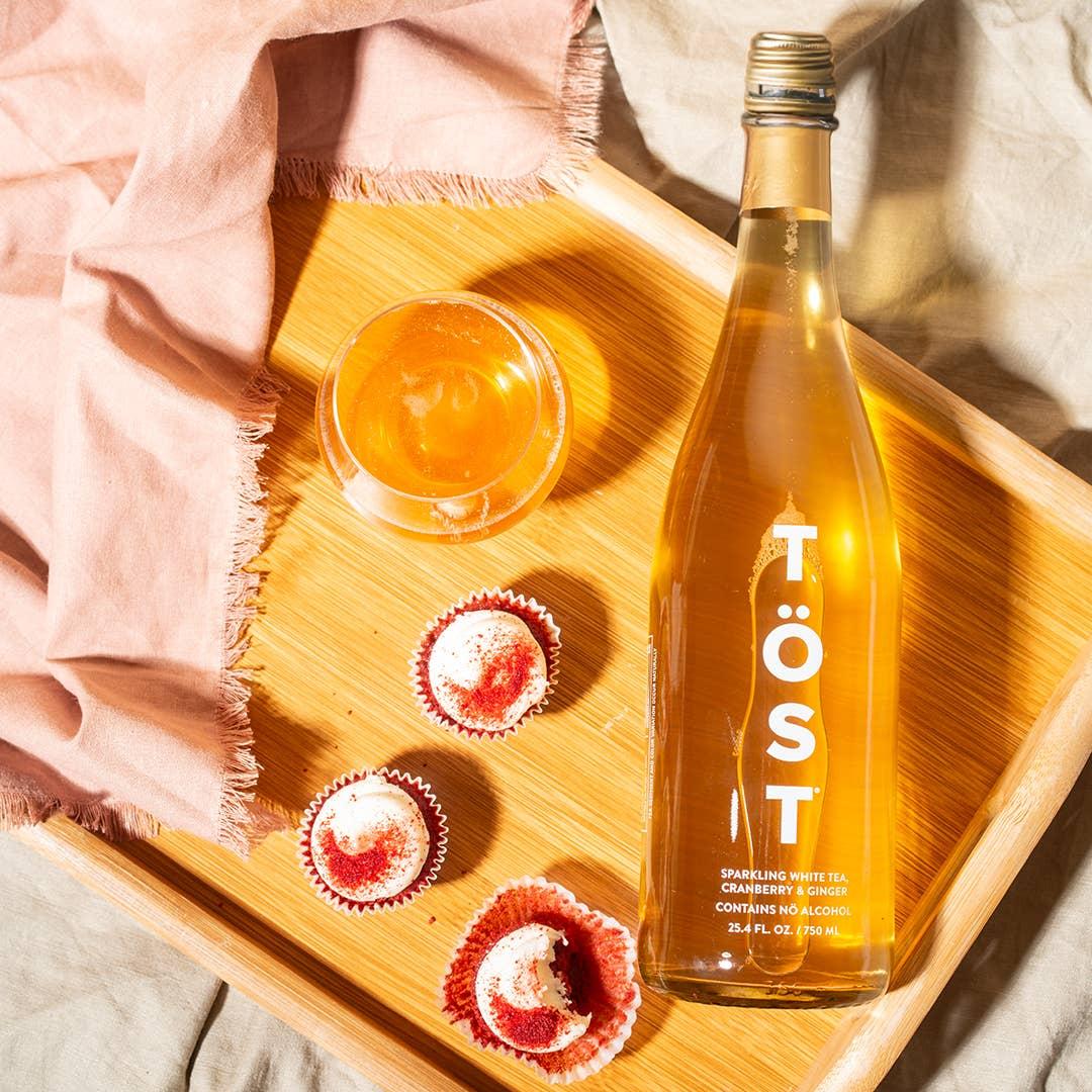 A bottle of TÖST a Non Alcoholic Refresher from TÖST Beverages, a refreshing non-alcoholic beverage, sits on a wooden tray accompanied by a filled glass and three red and white cupcakes. A pink cloth lies next to the items.