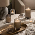 A cozy setting features the gentle glow of the Amelie- Linen Cotton Oasis 7oz Candle by La Jolie Muse, crafted from natural soy wax and housed in ribbed glass. This ambiance is heightened by a textured lamp and a decorative gold tray adorned with cotton branches, all enhanced by the subtle scent of white jasmine on a beige cloth.