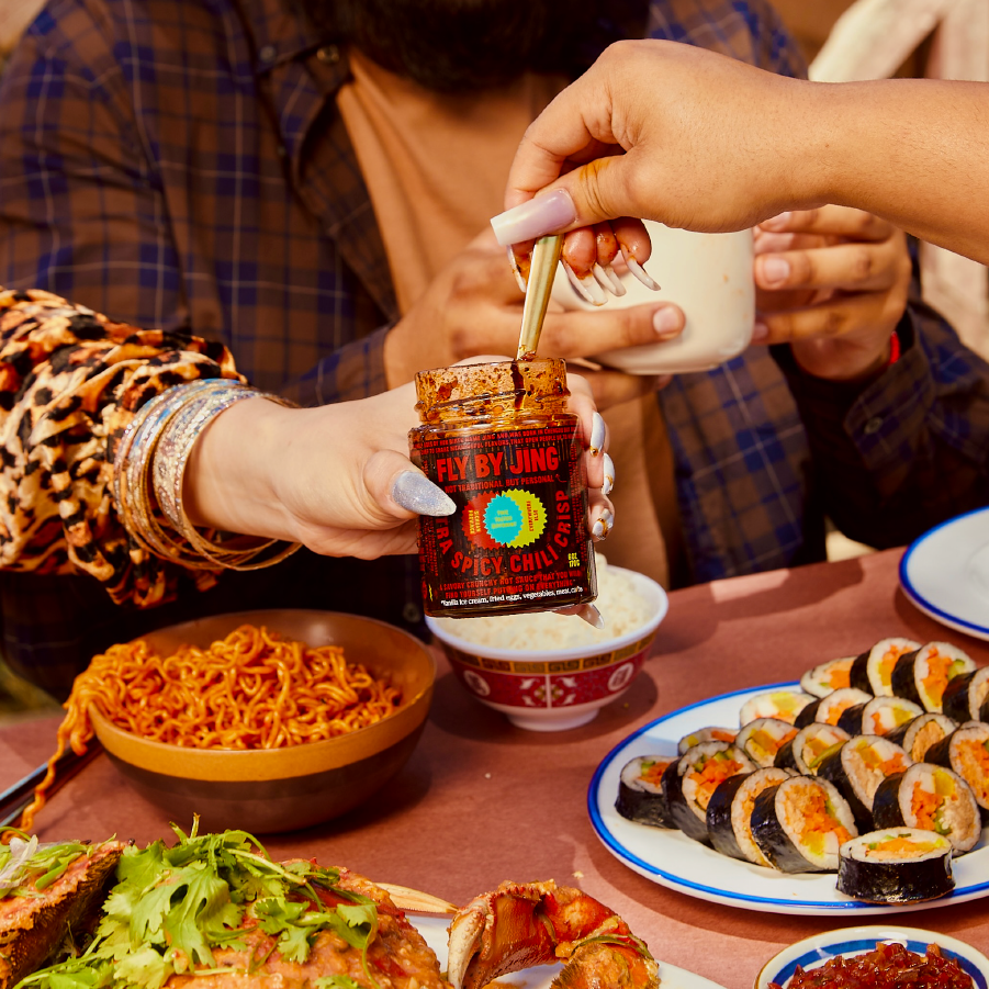 Two hands are holding a jar of Xtra Spicy Chili Crisp by Fly By Jing, hovering over a dining table filled with bowls of noodles, sushi, and various dishes.
