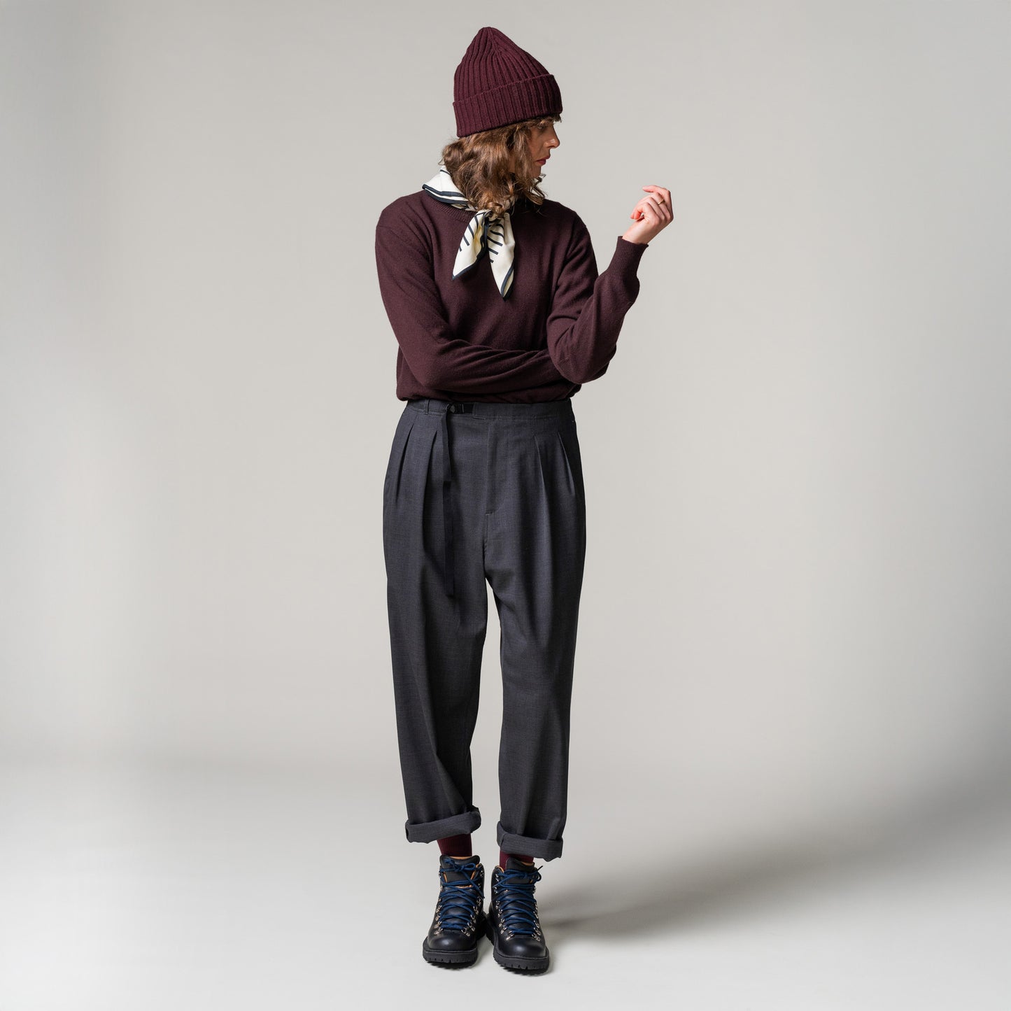 Dressed in a maroon Cashmere Crewneck Sweater by Seven Gauge, along with a beanie and scarf, gray cuffed pants, and black boots, the individual strikes a perfect pose against a plain backdrop, gazing to the side.