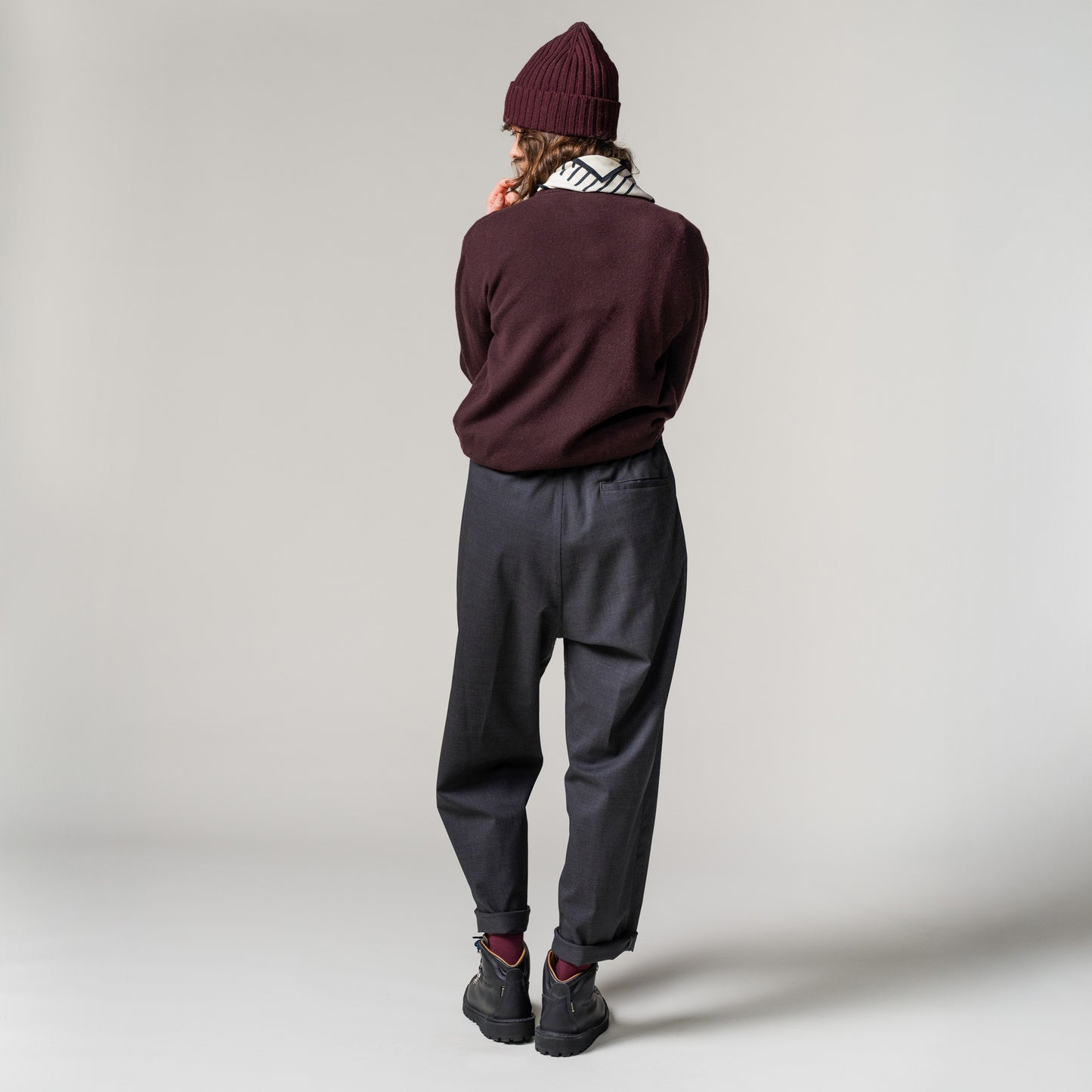 A person stands facing away, wearing a maroon beanie and a Seven Gauge Cashmere Crewneck Sweater. The look is completed with a striped cashmere scarf, gray pants, and black shoes, effortlessly blending into the plain gray background.